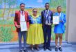 Four people stand in front of a colorful mural. Three students in distinct uniforms hold certificates and awards, smiling proudly. A teacher stands between them, wearing a vibrant dress. The scene conveys achievement and celebration.