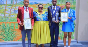 Four people stand in front of a colorful mural. Three students in distinct uniforms hold certificates and awards, smiling proudly. A teacher stands between them, wearing a vibrant dress. The scene conveys achievement and celebration.