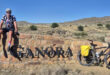 A cyclist in a colorful jersey sits on a stone wall with the word "Ganora" in front. A bike with panniers is nearby, set against a dry, hilly landscape.