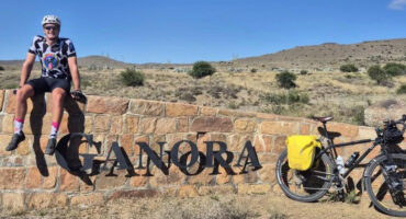 A cyclist in a colorful jersey sits on a stone wall with the word "Ganora" in front. A bike with panniers is nearby, set against a dry, hilly landscape.