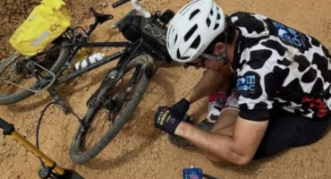 Cyclist on a dirt path fixes a flat tire on a black bike. He wears a white helmet and cow-print jersey. A yellow bag and pump lie nearby.