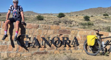 A cyclist in a patterned jersey and helmet sits smiling on a stone sign reading "GANORA." A bike with a yellow bag rests nearby. The landscape is arid with shrubs under a clear blue sky, creating an adventurous and cheerful vibe.