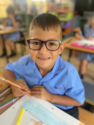 Young boy wearing glasses smiles while coloring at a classroom desk. The colorful environment and students in uniforms create a cheerful, focused mood.