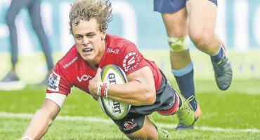 A rugby player in a red jersey dives across the grass clutching a ball, showing determination. Another player in blue shorts is in pursuit.