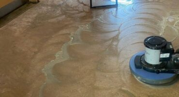 A floor cleaning machine polishes a wet, swirled floor in a hallway. A bench with bottles stands in the background. A worker cleans near the door.