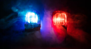 Red and blue police lights illuminate through swirling fog on a dark background, creating a dramatic and urgent atmosphere.