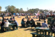 Outdoor fair with people seated at picnic tables, enjoying sunny weather. Green tractors and banners decorate the grassy area, creating a lively, community feel.