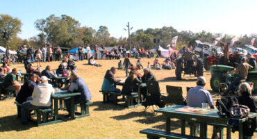 Outdoor fair with people seated at picnic tables, enjoying sunny weather. Green tractors and banners decorate the grassy area, creating a lively, community feel.