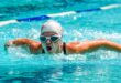 A swimmer in a white cap and goggles performs a butterfly stroke in a clear blue pool, exuding determination and focus, with water splashing energetically.
