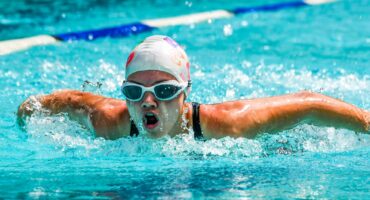 A swimmer in a white cap and goggles performs a butterfly stroke in a clear blue pool, exuding determination and focus, with water splashing energetically.