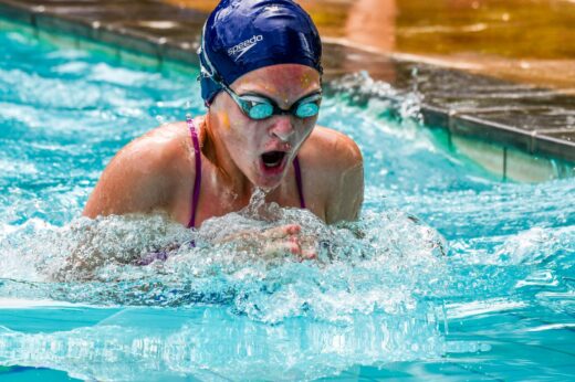 A swimmer in a blue cap and goggles competes in a pool, aggressively pushing through water. Her expression shows determination and focus.