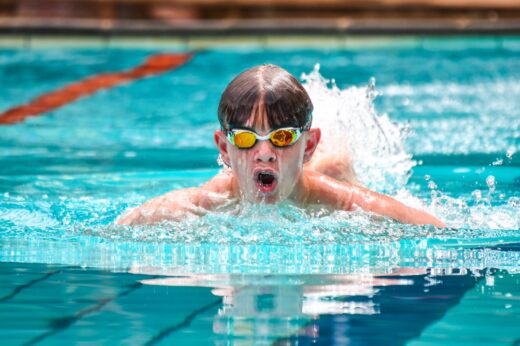 A swimmer in reflective goggles competes in a pool, face forward, creating splashes. The vibrant blue water and focused expression convey intensity.