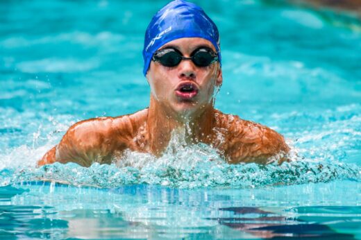 A swimmer in a blue cap and goggles powers through a pool, creating splashes. The focused expression and dynamic water convey determination.