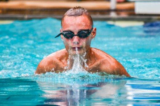 A swimmer wearing goggles emerges from a pool, splashing water around. The athlete appears focused and determined under the bright sunlight.