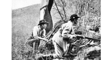 Two men in historical attire crouch on a rocky hillside, rifles in hand, appearing focused and alert. Sparse vegetation hints at a rugged, harsh landscape.