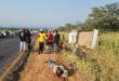 A group of people stands beside an overturned truck on a roadside, surrounded by grass. Emergency responders attend to a person lying on the ground. The scene is tense.