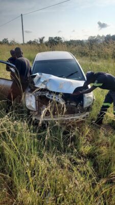 A damaged car with a crumpled front is parked in tall grass. Three people are inspecting the car, conveying concern and focus under a partly cloudy sky.