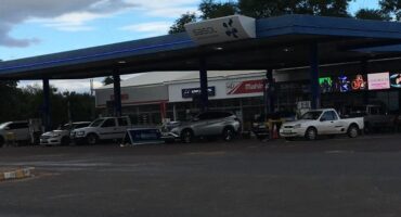 Gas station scene with multiple parked cars under a blue canopy. The signage reads “Sasol." Overcast sky and trees frame the background.