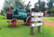 Antique tractor with rusted orange wheels on a lush green field, next to directional signs for a museum, picnic area, and homestead, evoking nostalgia.