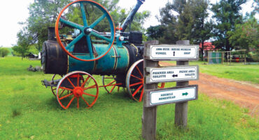 Antique tractor with rusted orange wheels on a lush green field, next to directional signs for a museum, picnic area, and homestead, evoking nostalgia.