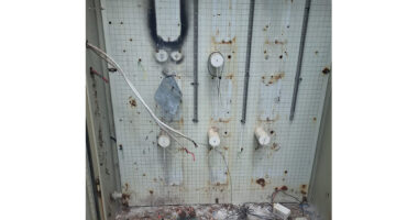 Rusty electrical panel with wires and components removed, set against a gridded metal backdrop. Weathered and abandoned, suggesting neglect and disuse.