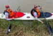 Two people in a long white kayak smiling, wearing orange life vests and caps, paddle by grassy riverbank. The atmosphere is cheerful and adventurous.