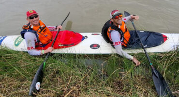 Two people in a long white kayak smiling, wearing orange life vests and caps, paddle by grassy riverbank. The atmosphere is cheerful and adventurous.