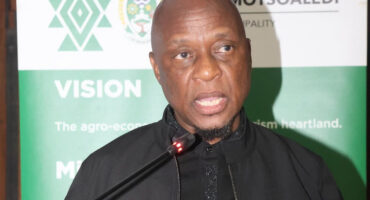 A person speaks at a podium with a microphone, against a green and white backdrop featuring text and logos. The tone appears formal and professional.