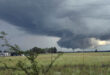 A vast field under a dramatic, dark gray cloud formation suggesting a potential thunderstorm and tornado. A faint green landscape contrasts with the ominous sky.