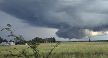 A vast field under a dramatic, dark gray cloud formation suggesting a potential thunderstorm and tornado. A faint green landscape contrasts with the ominous sky.