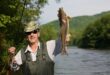 A person wearing sunglasses, a camo hat, and fishing gear holds up a large fish triumphantly by a scenic river, surrounded by lush greenery.