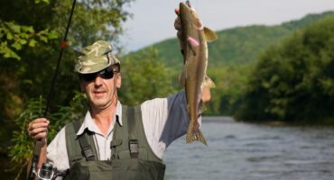 A person wearing sunglasses, a camo hat, and fishing gear holds up a large fish triumphantly by a scenic river, surrounded by lush greenery.