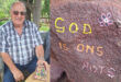 A smiling man in a plaid shirt sits outdoors, displaying a painted rock. Next to him, a rock has the message "GOD IS ONS ROTS" with painted flowers. The setting is calm and natural.