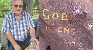 A smiling man in a plaid shirt sits outdoors, displaying a painted rock. Next to him, a rock has the message "GOD IS ONS ROTS" with painted flowers. The setting is calm and natural.