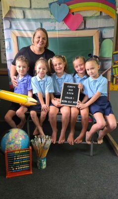A teacher and five smiling children in uniforms sit on a bench. There's a globe, paintbrushes, abacus, and a sign reading their first day of grade one.