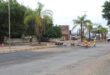 Street work scene with palm trees lining the road. Traffic cones and caution signs surround a repair area. A parked vehicle and overcast sky in the background.