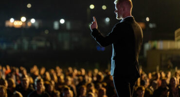 A man in a suit stands on stage, gesturing with one finger raised, addressing a large, attentive crowd under soft lighting at night.