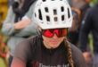 A female cyclist wearing a white helmet and red sunglasses focuses with determination. She's in a black jersey with "Tipping Point" text. Blurred spectators in the background.