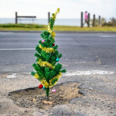 A small Christmas tree, adorned with colorful ornaments and gold tinsel, is humorously planted in a road pothole. A blurred seaside landscape is in the background.