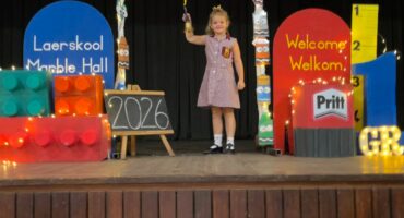 A young girl in a checkered school uniform stands on a decorated stage holding a torch. Surrounding her are colorful displays, a chalkboard with "2026," and a welcome sign, creating a festive, welcoming atmosphere.