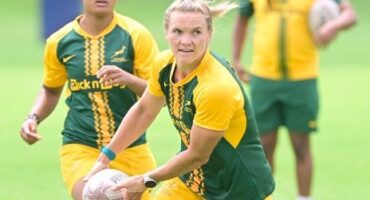 A woman rugby player in a green and yellow uniform holds the ball, looking focused. Teammates in similar kits are in the background on a field.