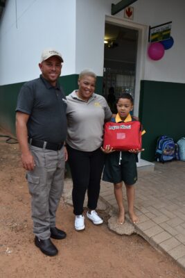 A smiling man and woman stand beside a child holding a red item labeled with "Logan Nkosi" outside a classroom. School bags are visible in the background.