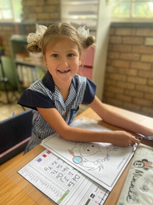 Young girl in a school uniform smiles at a desk, working on a coloring sheet. Sunlight streams through a window, creating a warm, cheerful atmosphere.