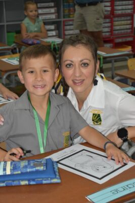 A young boy and a woman sit at a classroom desk, smiling at the camera. The boy holds a marker, and school supplies and worksheets are on the desk. The mood is cheerful and welcoming.