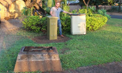 A person stands on a green lawn near two utility boxes, one green and one beige, under a tree, in front of a residential area, smiling confidently.