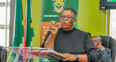 A woman in a black dress and glasses speaks passionately at a podium. Behind her are flags and a green banner, conveying an official setting.