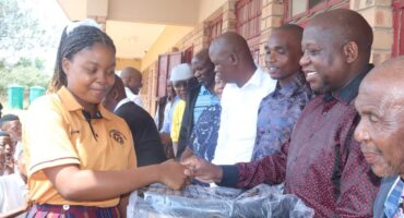 A young woman in a school uniform smiles as she shakes hands with a smiling man. They stand in front of a group of people, suggesting a celebratory event.
