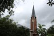A tall brick clock tower with a pointed roof stands amidst lush green trees. The sky is overcast, suggesting an impending storm, creating a somber tone.