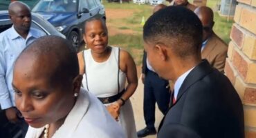 A group of people in formal attire stand and converse near a vehicle and brick wall. The mood appears serious yet cordial.