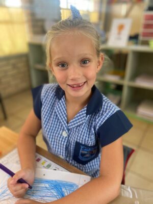 Smiling young girl in a blue checkered school uniform draws with a purple marker. She's seated at a classroom table with bookshelves in the background.